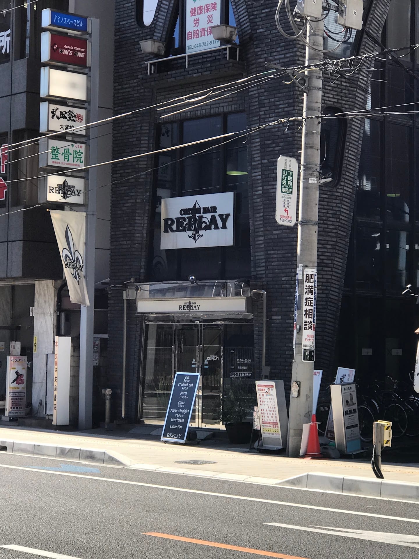 黒いタイル張りのビルの入口にREPLAYの看板が掲げられ、ガラス張りのドアの前にポスターが並んでいる